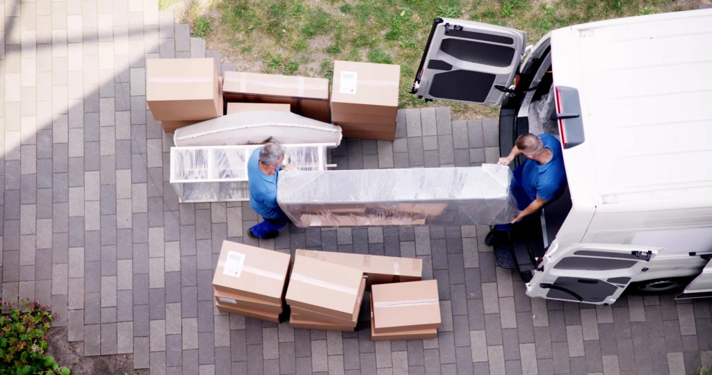 Zwei Männer entladen verpackte Kisten und Neumöbel aus einem Lastwagen eines Umzugsunternehmens.